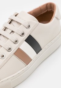 Baskets beige clair à bout arrondi, arborant des rayures contrastées en noir, blanc et beige. Partie supérieure en cuir lisse, design à lacets, semelle en caoutchouc.