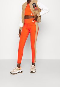 Orange atletisk cropp-topp och hög midja leggings med beige sidremsor, i kombination med en tan och vit letterman-jacket och sneakers.