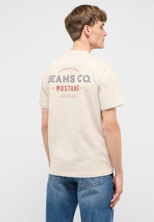 Young man wearing beige short-sleeve shirt with "Jeans Co. Mustang" text on the back, paired with blue denim jeans, facing sideways.