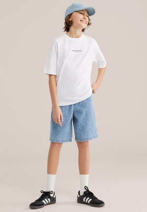 Naeratav laps kannab helesinist nokamütsi, valget teksti sisaldavat t-särki, teksapükse, valgeid sokke ja musti Adidas tossusid, seisab käsi puusal.