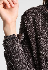 Grå, plysch-fleeceplagg med en texturerad yta och elastiska manschetter, med ett mjukt, fluffigt utseende och subtil glans.