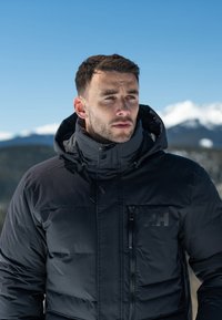 Black insulated jacket with a high collar, zippered pockets, and logo on chest. Smooth texture, designed for cold weather.