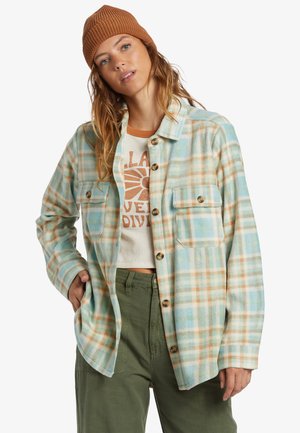 Flannel shirt in light blue with orange and cream plaid pattern, featuring two chest pockets and brown buttons, paired with olive green pants.