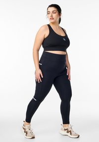 Czarny stanik sportowy oraz czarne legginsy z wysokim stanem i kieszeniami. Legginsy mają subtelne odblaskowe akcenty; model nosi beżowo-brązowe sneakersy.