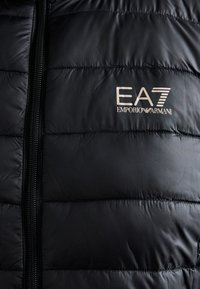 Crna jakna s podstavom i zatvaračem, koja sadrži vertikalne šavove i srebrni EA7 Emporio Armani logo na prsima. Glatka tekstura.