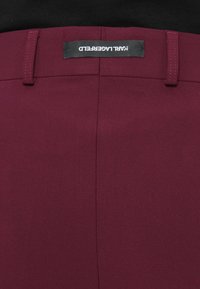 Bordo kelnės su lygia tekstūra, turinčios juosmens etiketę su užrašu "KARL LAGERFELD." Švarus dizainas be matomų raštų ar detalių.