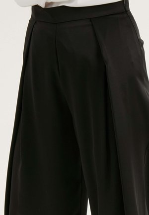 Pantalons noirs ajustés avec une texture lisse, des plis structurés à l'avant et une large taille.