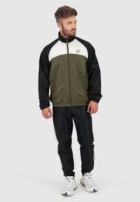 Giacca a zip verde e nera con pannello bianco sul petto, polsini elasticizzati, abbinata a pantaloni da tuta neri con strisce bianche ai lati.