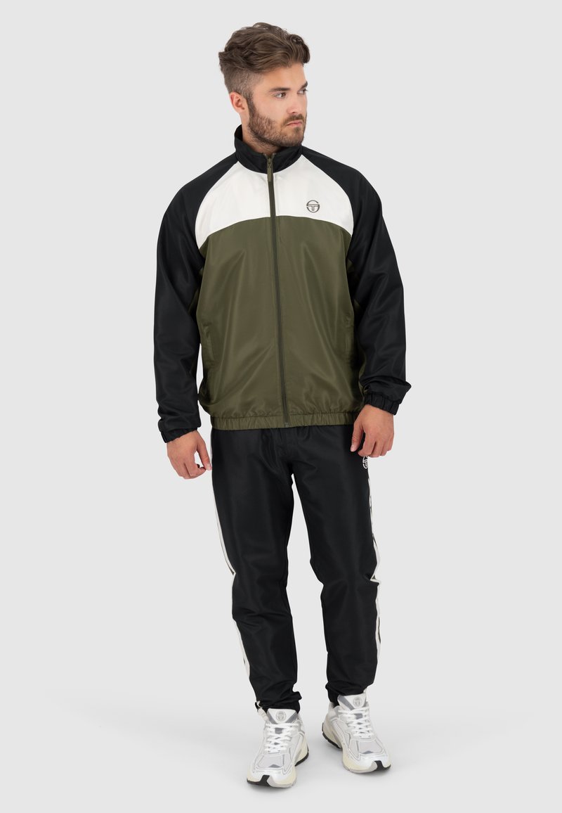 Giacca a zip verde e nera con pannello bianco sul petto, polsini elasticizzati, abbinata a pantaloni da tuta neri con strisce bianche ai lati.