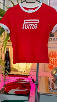 Red Puma short-sleeve t-shirt with white accents and logo. Soft fabric, cropped design, displayed on a hanger with plants in background.