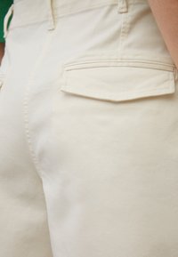 Beige kangas-housut, joissa on sileä pinta, slim fit -malli ja takataskut viistolla läpällä. Ompeleet parantavat muotoilua.