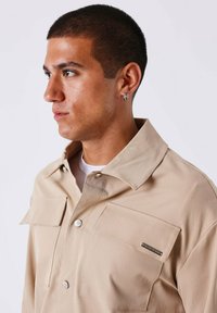 Camicia beige con bottoni, dal colletto ampio, due tasche frontali e un accento di branding discreto. Il tessuto appare liscio con una vestibilità strutturata.