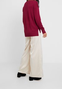 Burgunderfarbener Strickpullover mit seitlichen Schlitzen, kombiniert mit cremefarbenen, weit geschnittenen Hosen aus glattem Stoff. Schwarze Schuhe runden das Outfit ab.