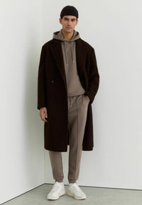 Hosszú barna kabát bézs kapucnis pulóverrel és hozzáillő, szűk szárú nadrággal. Fehér tornacipők emelik az összhatást. Egyszerű háttér előtt.