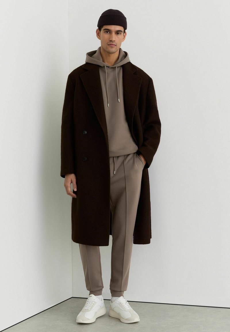 Man wearing a taupe hoodie and matching joggers beneath a long dark brown coat with white sneakers