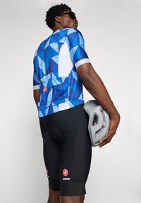 Maillot de cyclisme à motif géométrique bleu avec des manches courtes, associé à un short rembourré noir ; tenant un casque gris.
