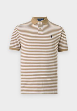 Jeune homme debout, portant un polo à rayures beige et blanche, un jean bleu clair et des baskets blanches sur fond blanc uni.
