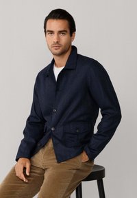 Marineblauwe wollen blend jasje met een kraag, knoopsluiting en twee voorzakken met flap. Draagt over een wit overhemd en corduroy broek.