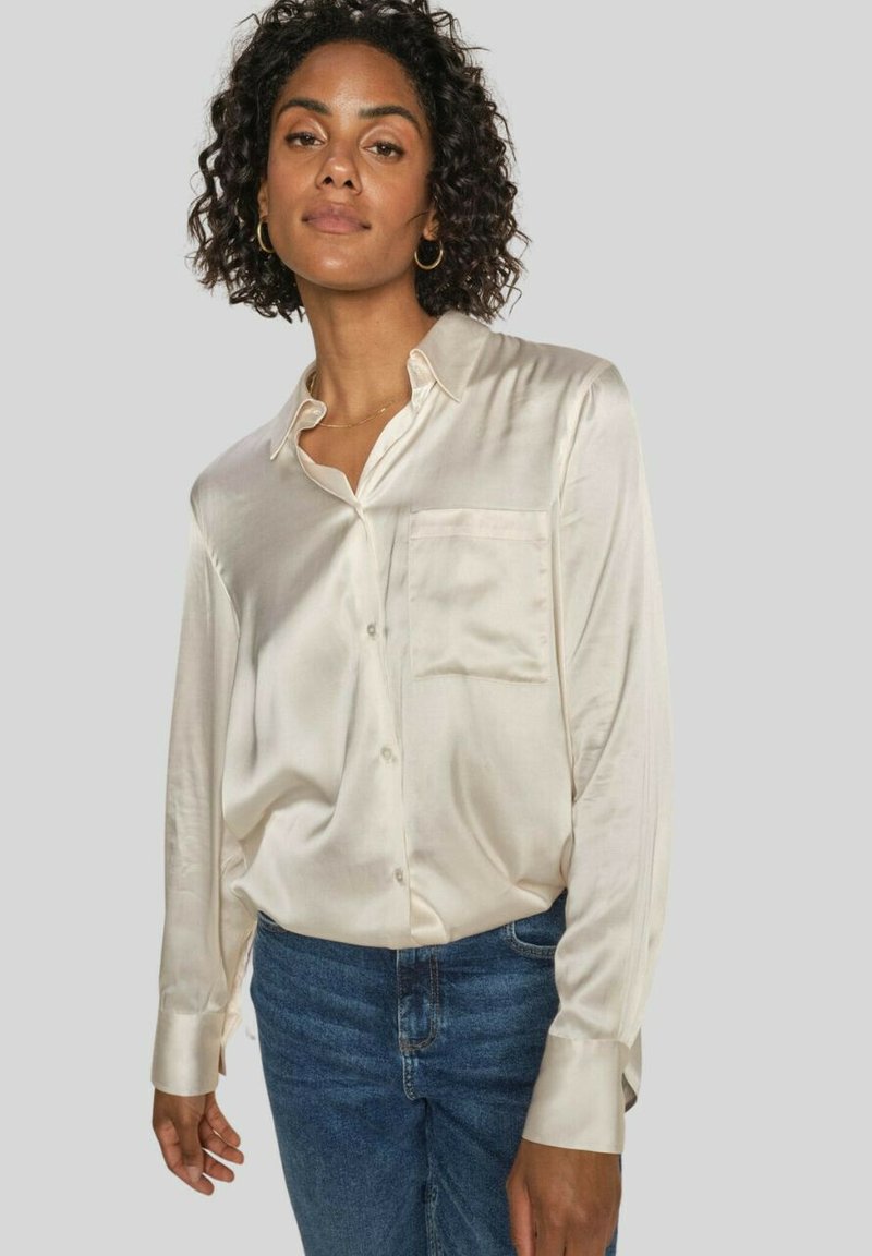 Cream satin button-up shirt with a chest pocket, long sleeves, and a relaxed fit, paired with blue denim jeans.