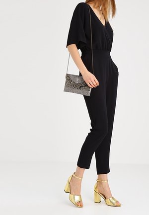 Femme portant une combinaison noire, tenant un sac bandoulière argenté texturé, et des sandales à talons blocs dorés, debout devant un fond uni.