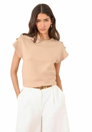 Ung kvinde med langt mørkt hår iført en beige top med korte ærmer og hvide bukser med høj talje, med hænderne i lommerne.