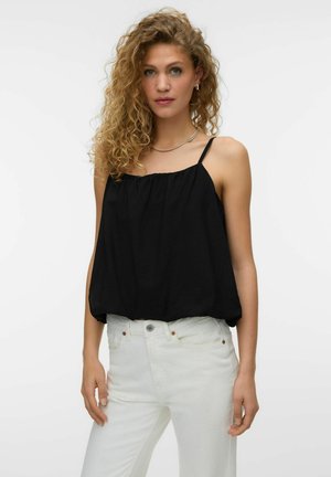 Frau mit lockigem blonden Haar, die ein schwarzes ärmelloses Oberteil und weiße Jeans trägt, vor einem schlichten hellen Hintergrund stehend.