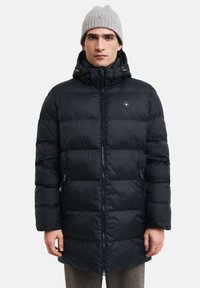 Sort pufferjakke med lynlås i front, en aftagelig hætte og et rundt logo på brystet. Har lynlåslommer og en quiltet tekstur.