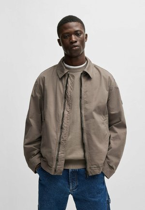 Light brown bomber jacket with a zip closure, collared neck, and side pockets, layered over a beige sweater, paired with blue denim cargo pants.