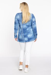 Blue denim shirt with a textured print, featuring a button-down back and long sleeves with cutout accents, paired with white zippered trousers.
