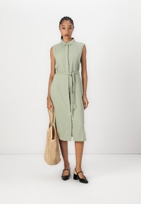 Light green, sleeveless dress with a collar and belt, made of textured fabric. Worn with black shoes and holding a woven bag.