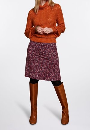 Kvinde iført en orange strikket sweater, knælængde nederdel med blomsterprint, sorte strømpebukser og brune knæhøje læderstøvler står foran en hvid baggrund.
