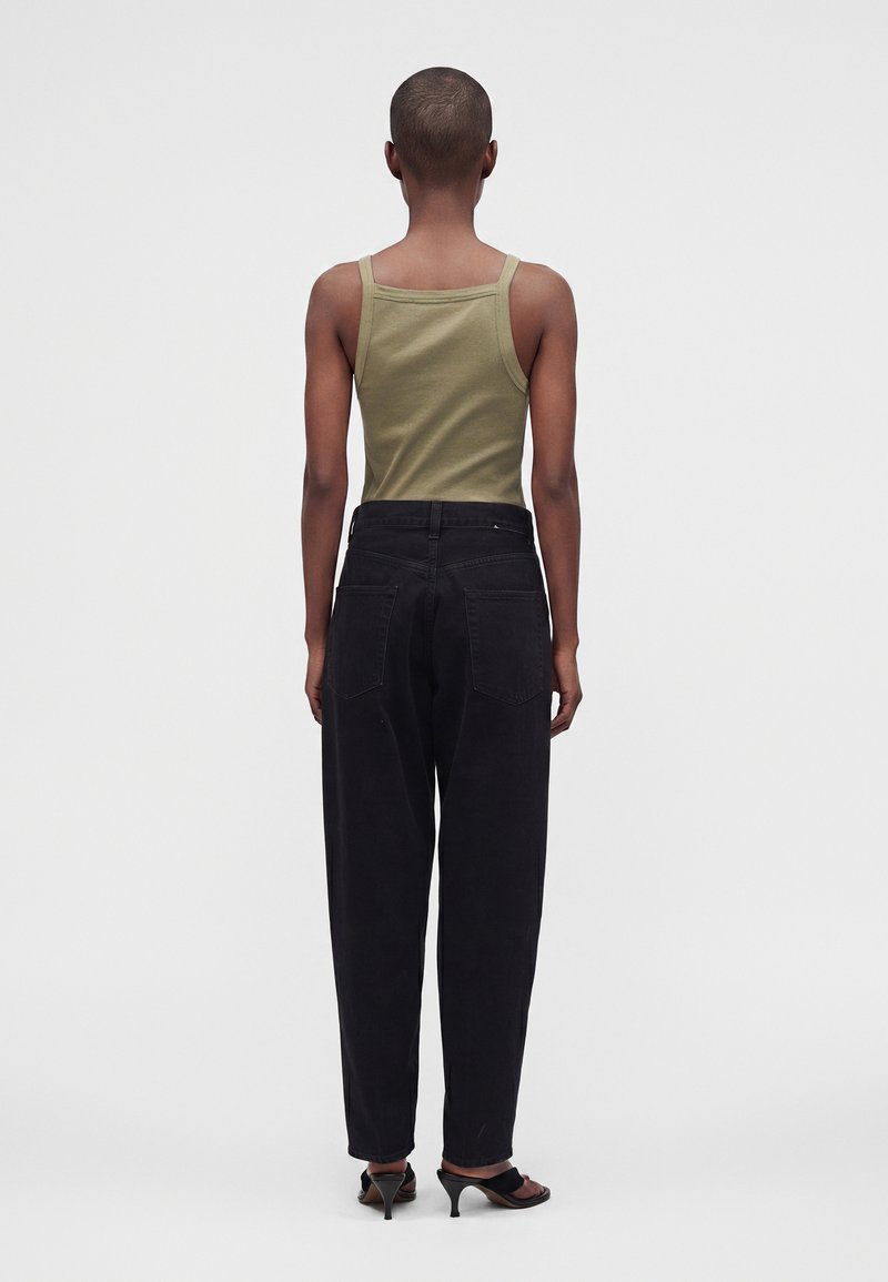 Personne aux cheveux courts portant un débardeur vert olive, un pantalon ample noir et des sandales à talons noires, debout de dos sur un fond uni.