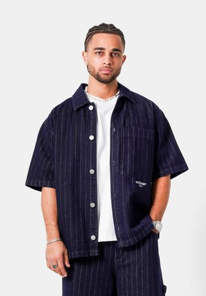 Mand med flettet hår iført navy blå pinstripe kortærmet skjorte, hvid T-shirt, matchende bukser, sølvur, halskæde og armbånd mod hvid baggrund.
