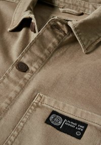 Beige knapp-up skjorta i bomullsblandning med en bröstficka. Har metallknappar och en svart etikett med vit text. Slät textur.