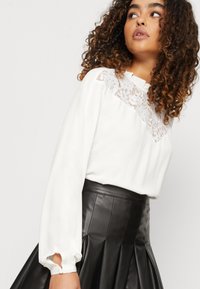 Hvit bluse med blonde detaljer ved halslinjen, med lange ermer med rynkede mansjetter. Kombinert med et svart skinnskjørt med folder.