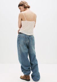 Kvinna med kort hår som bär en genomskinlig vit linne, lösa blå jeans och bruna ankelboots, poserar med ryggen vänd och tittar ner.