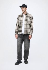 Koszula w kratę w odcieniach beżu i czerni, zestawiona z białym t-shirtem i szarymi dżinsami. Czarna czapka i ciemne trampki dopełniają zestaw.