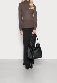 Femme en col roulé marron et pantalon noir tenant un grand sac à main en cuir noir, debout devant un fond blanc uni.