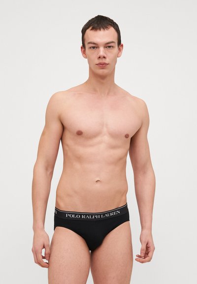 Μαύρο σλιπάκι με ελαστική μέση με εκτύπωση λογότυπου. Το ύφασμα φαίνεται λείο μ�ε εφαρμογή που αγκαλιάζει καλά, σε αντίθεση με τον τόνο του δέρματος.