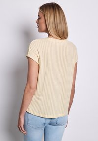 Světle žlutý žebrovaný top s krátkým rukávem a kulatým výstřihem, s uvolněným střihem a měkkou texturou, v kombinaci se světle modrými džínami.