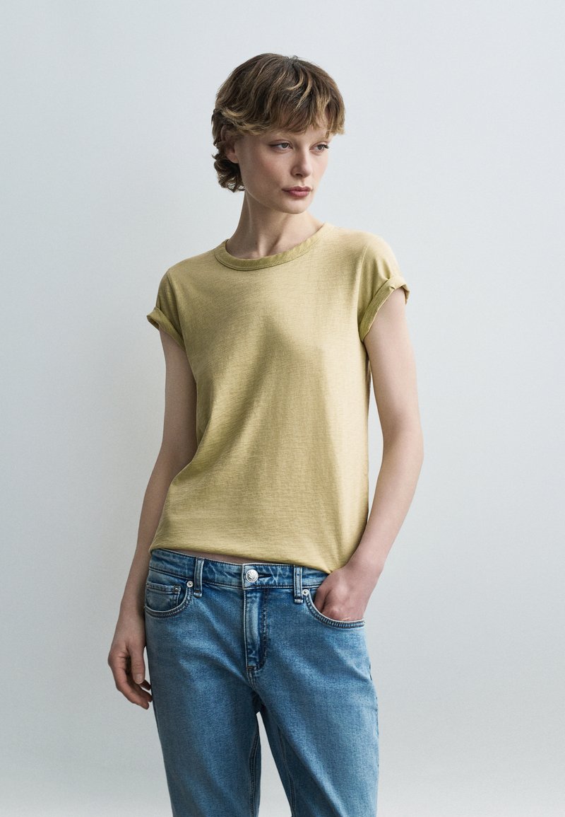 Νεαρός ενήλικας με κοντά μαλλιά φοράει κίτρινο κοντομάνικο μπλουζάκι και μπλε τζιν, στέκεται με το ένα χέρι στην τσέπη μπροστά σε απλό φόντο.