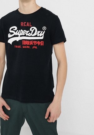 Osoba ubrana w czarną koszulkę SuperDry z czerwonym i białym napisem, w parze z ciemnozielonymi spodniami, stojąca na jednolitym jasnym tle.