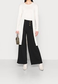 Bílý dlouhý kardigan přes světlejší top, kombinovaný s černými širokými kalhotami s třemi zlatými knoflíky. Béžové kotníkové boty a malá taška dotvářejí vzhled.