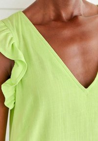 Blouse vert citron avec un col en V et des manches à volants. Le tissu a une finition texturée avec un motif à carreaux subtil.