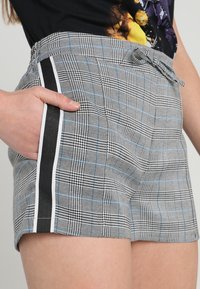 Gros plan d'une personne portant un short à carreaux gris avec des lignes bleues et des rayures noires sur les côtés, la main reposant dans la poche, associé à un haut graphique noir.