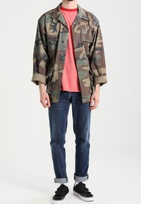 Chaqueta de camuflaje hecha de una tela resistente, combinada con una camiseta roja. Unos jeans azules con los puños doblados y unas zapatillas negras completan el atuendo.