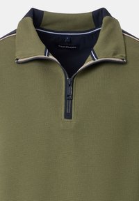 Grön zip-neckt tröja med ribbad textur, kontrasterande marinblå detaljer på kragen och en vävd etikett. Har en halv längd dragkedja.