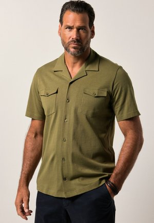 Man met kort donker haar en baard, die een olijfgroen overhemd met korte mouwen en borstzakken draagt, tegen een effen achtergrond.
