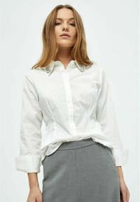 Camicia bianca con colletto decorato e lucido, maniche lunghe e vestibilità sartoriale, abbinata a una gonna grigia in un tessuto testurizzato.