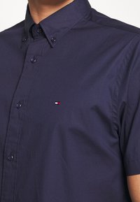Primo piano di una persona che indossa una camicia a maniche corte blu navy con bottoni, con un piccolo logo rosso, bianco e blu sul petto.